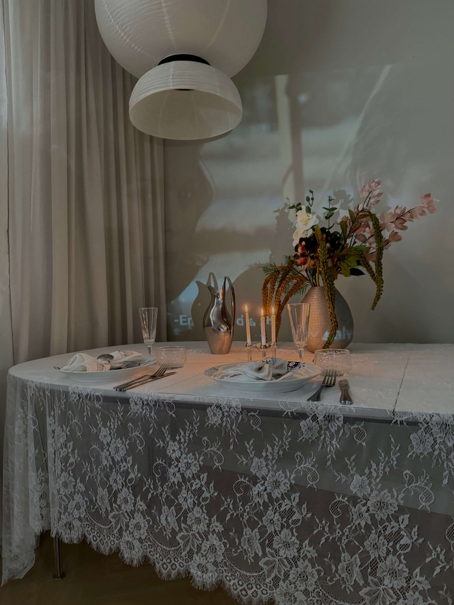 Hvid blondedug i en stemningsfuld aftenopsætning med levende lys, blomster og et dækket spisebord. Skaber en hyggelig og romantisk atmosfære.