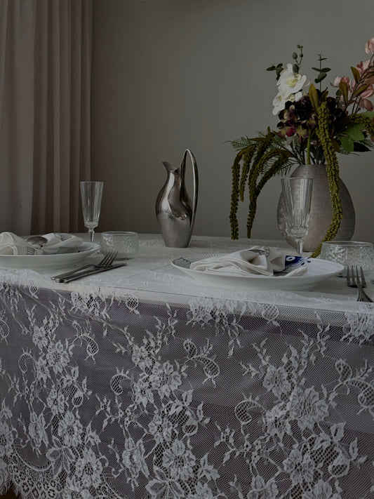 En smuk hvid blondedug på et opdækket spisebord med blomster, glas og en elegant sølvkande. Den gennemsigtige dug fremhæver bordets naturlige skønhed.