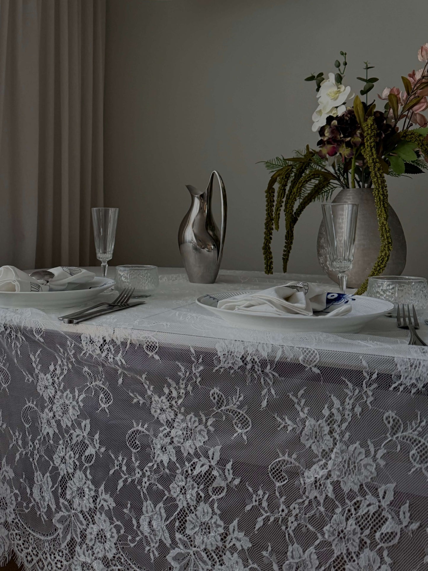 En smuk hvid blondedug på et opdækket spisebord med blomster, glas og en elegant sølvkande. Den gennemsigtige dug fremhæver bordets naturlige skønhed.
