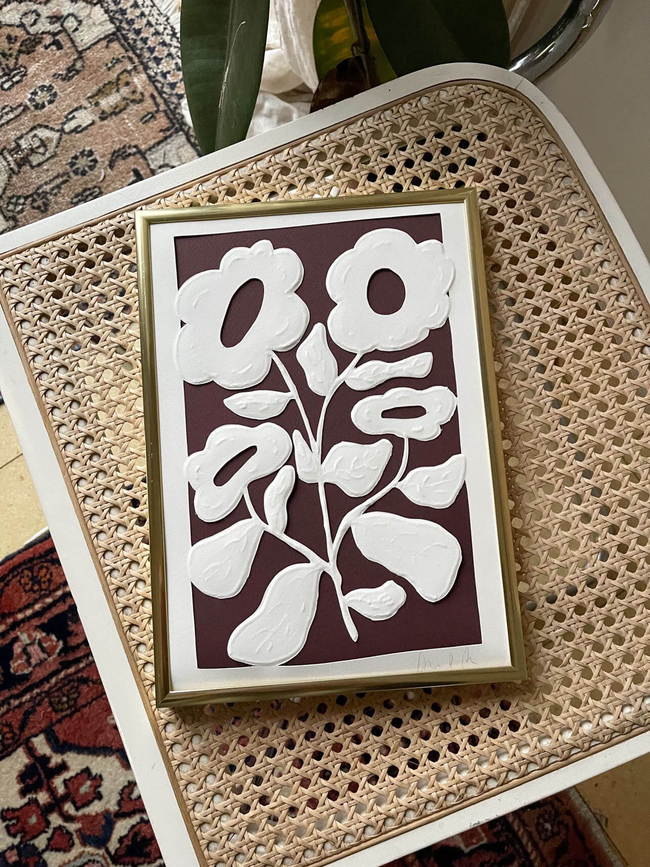 Blomster linoleumstryk i messingramme, placeret på stol med flet – håndlavet kunst fra Camilla Bergqvist Andersen.