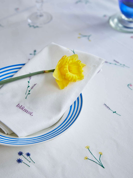 Hvid stofserviet med broderede vilde blomster, foldet på en tallerken med en gul blomst ovenpå.