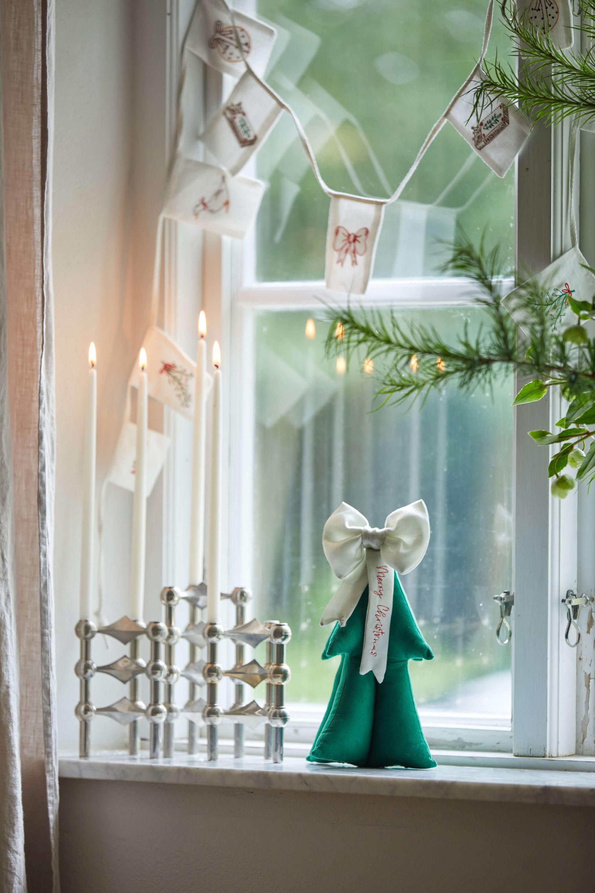 Stemningsfuld guirlande med broderi hænger i et vindue med et juletræ i baggrunden