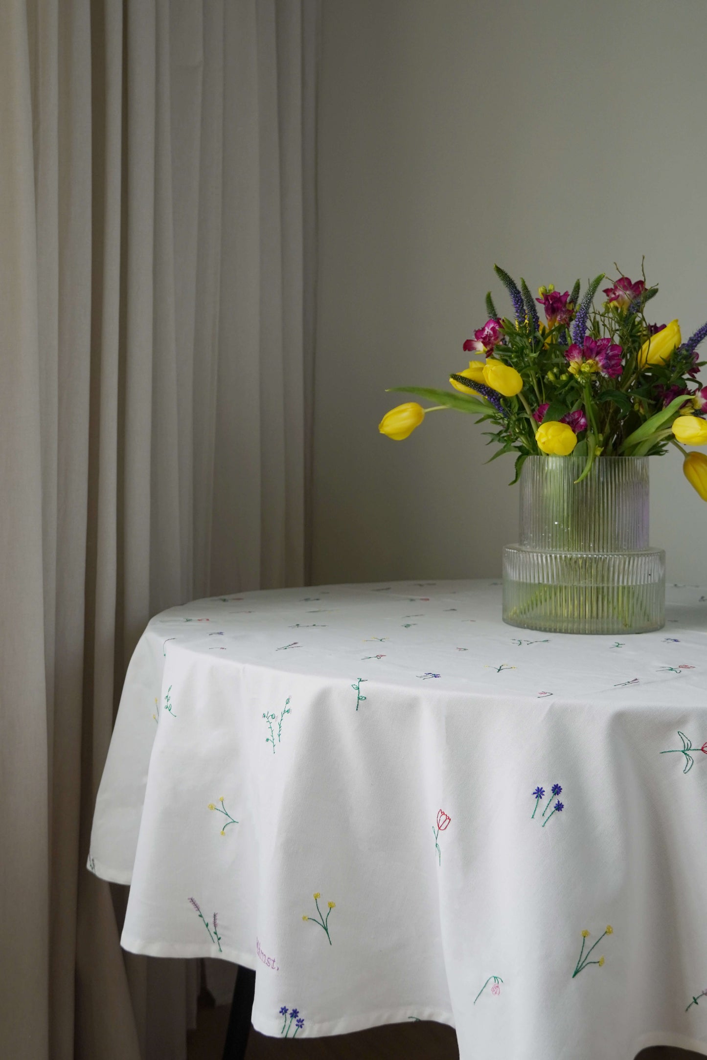 Rundt bord med hvid dug broderet med blomster, pyntet med en vase med farverige blomster, foran lyse gardiner.