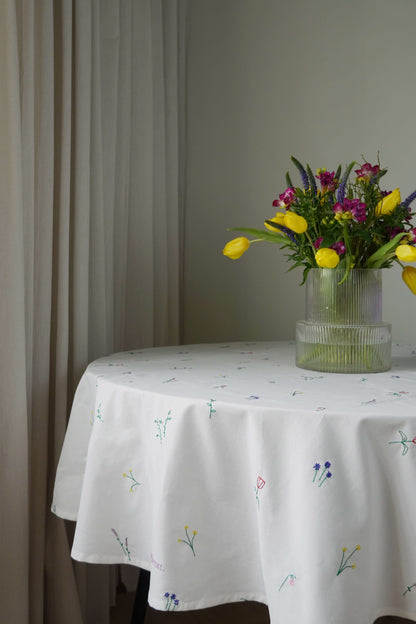 Rundt bord med hvid dug broderet med blomster, pyntet med en vase med farverige blomster, foran lyse gardiner.