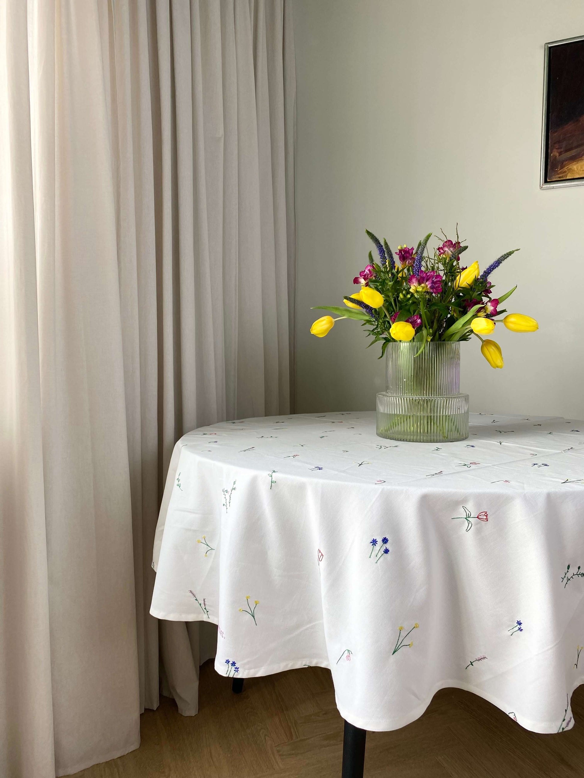 pisebord dækket med en hvid broderet dug og en vase med blomster, placeret i et hjørne af et rum med beige gardiner.