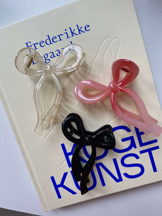 Tre Bow Claw hårklemmer i sort, rosa og transparent, placeret på en bog – stilfuldt design til enhver anledning.