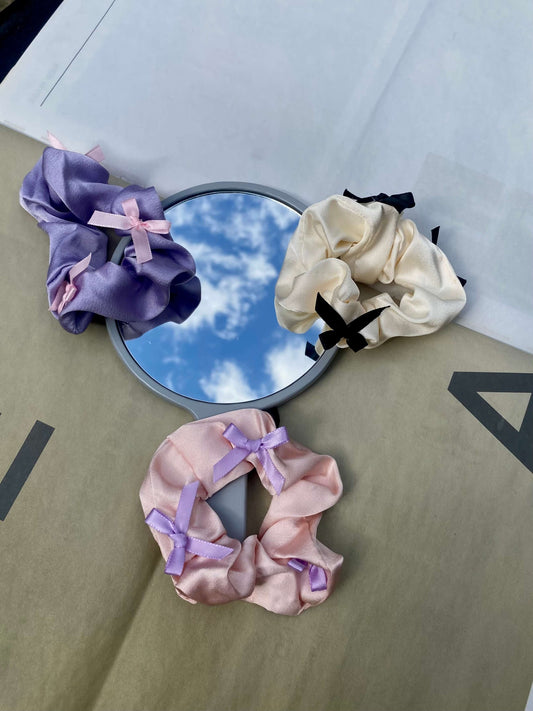 Bow Scrunchies i lilla, lyserød og creme, præsenteret på et spejl med himmelrefleksion.