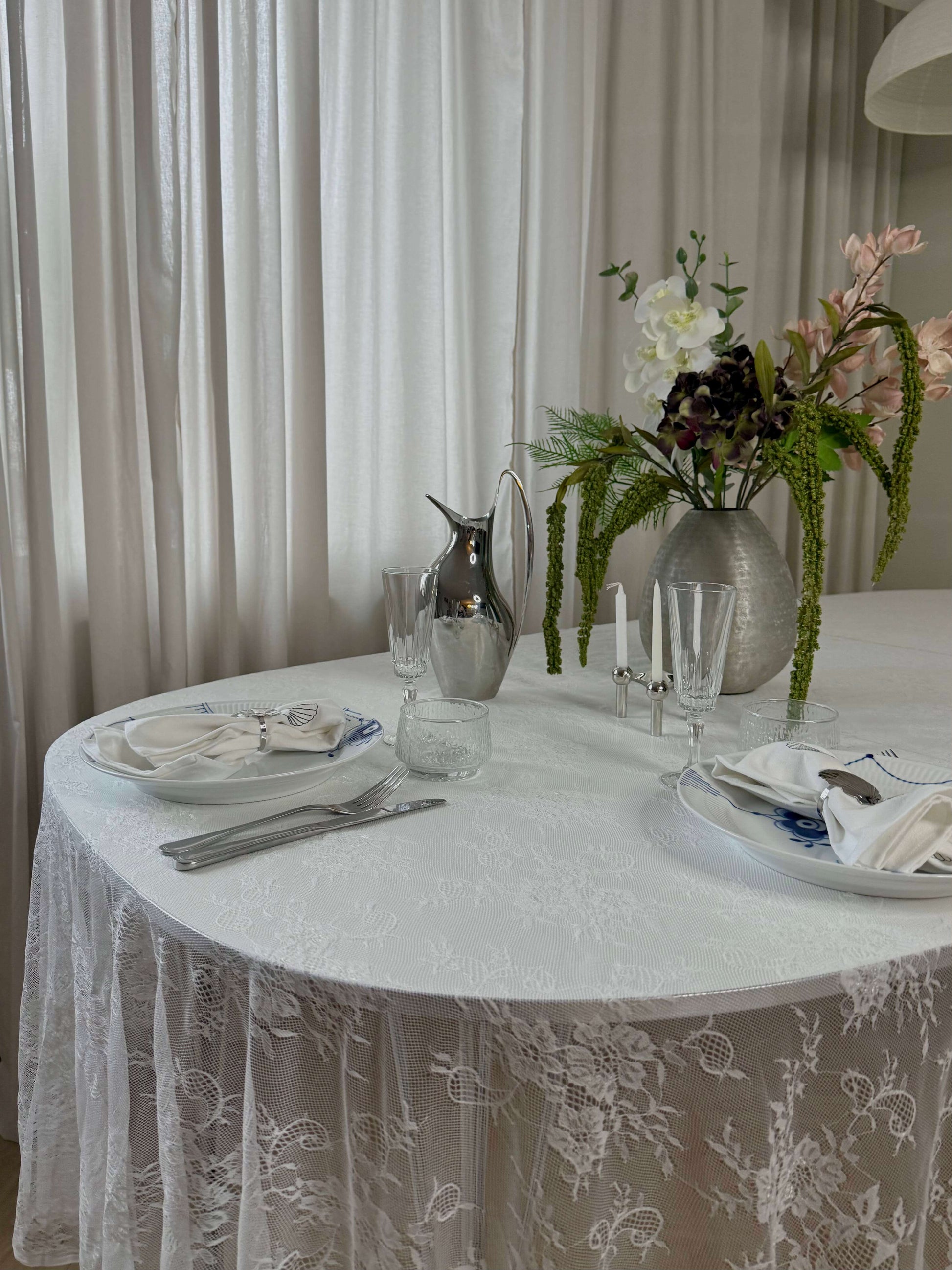 Hvid blondedug på et rundt bord med blomster og sølvkande. De gennemsigtige blonder giver et let og luftigt look til borddækningen.