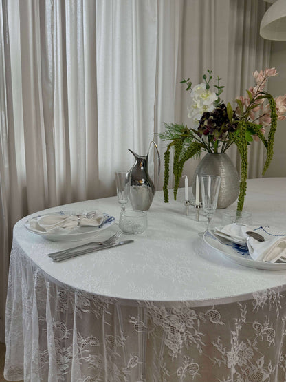 Hvid blondedug på et rundt bord med blomster og sølvkande. De gennemsigtige blonder giver et let og luftigt look til borddækningen.