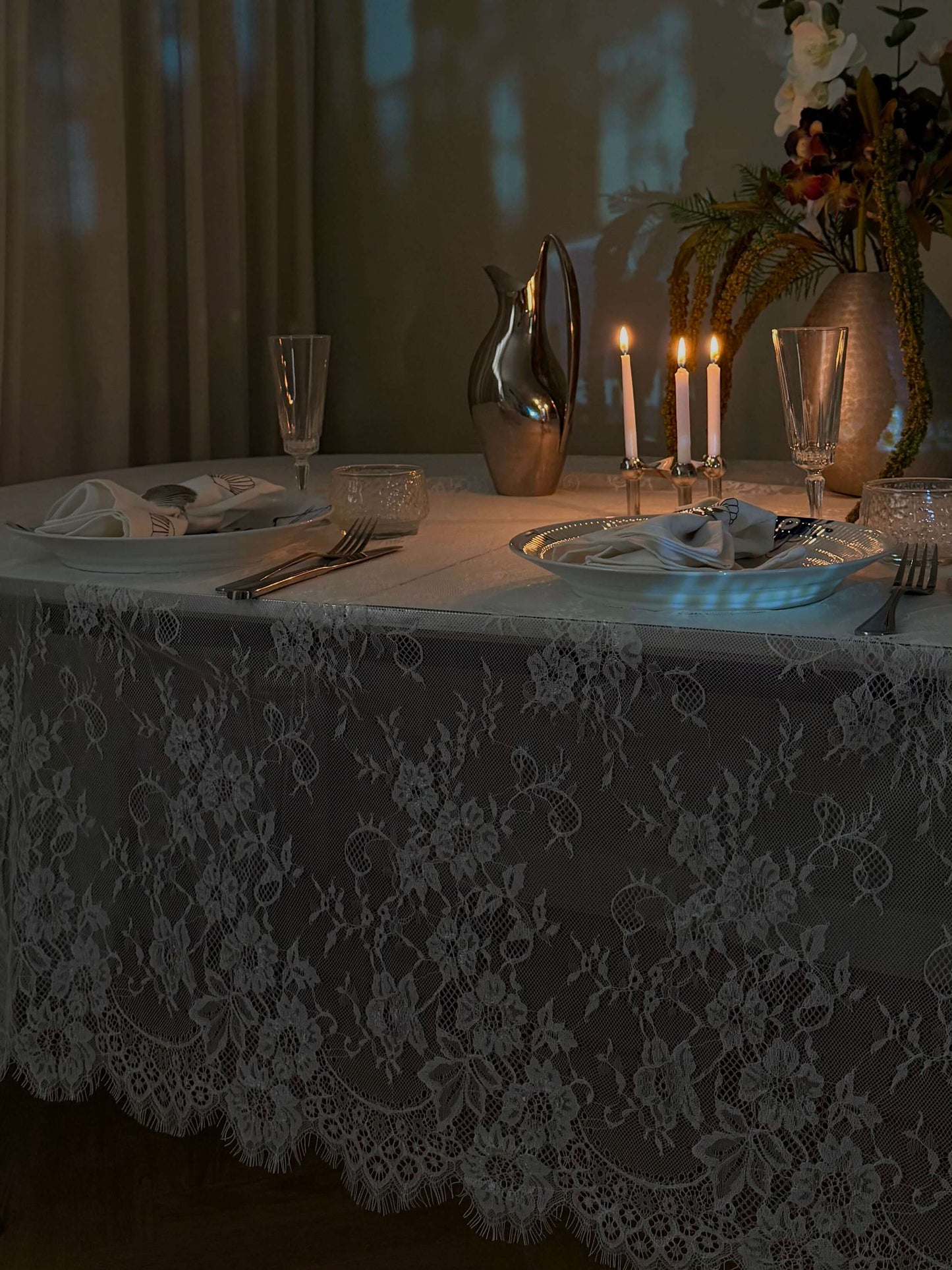 Hvid blondedug på et spisebord i aftenbelysning med levende lys, glas og blomsterdekorationer. Perfekt til en romantisk middag.