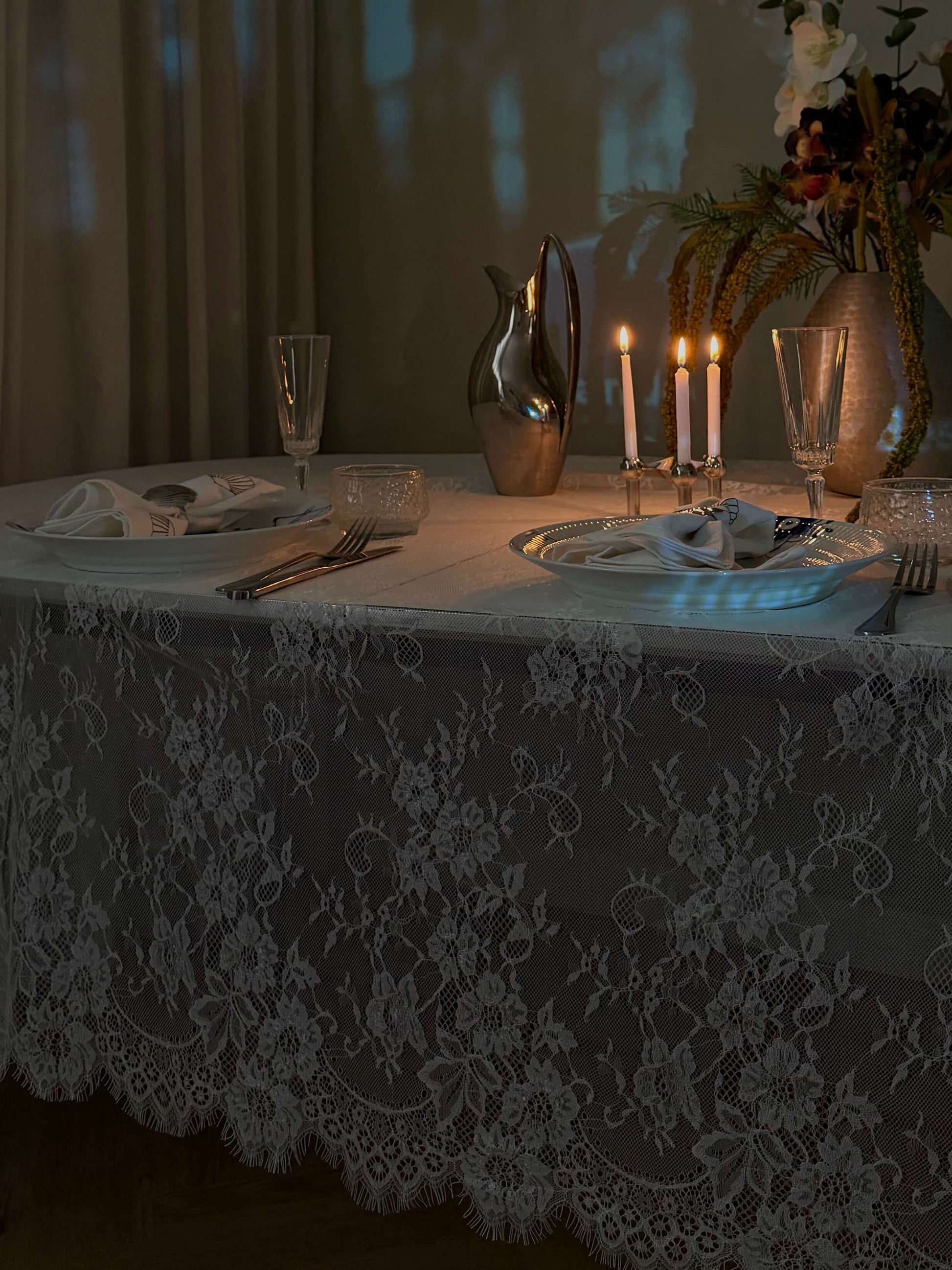 Hvid blondedug på et spisebord i aftenbelysning med levende lys, glas og blomsterdekorationer. Perfekt til en romantisk middag.