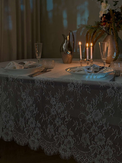Hvid blondedug på et spisebord i aftenbelysning med levende lys, glas og blomsterdekorationer. Perfekt til en romantisk middag.