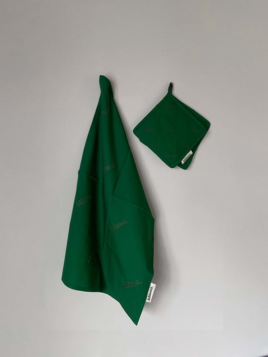 Grøn grydelap fra Blåmst hængende på væggen, lavet af økologisk bomuld med broderet logo. Enkel og stilren køkkendetalje.
