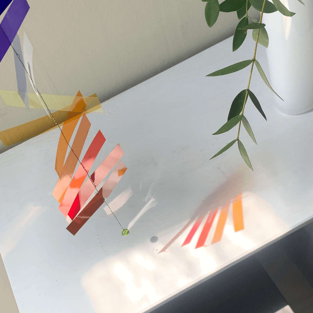 Detaljebillede af Flickering Light uro med sollys, der skaber refleksioner på en hvid flade.