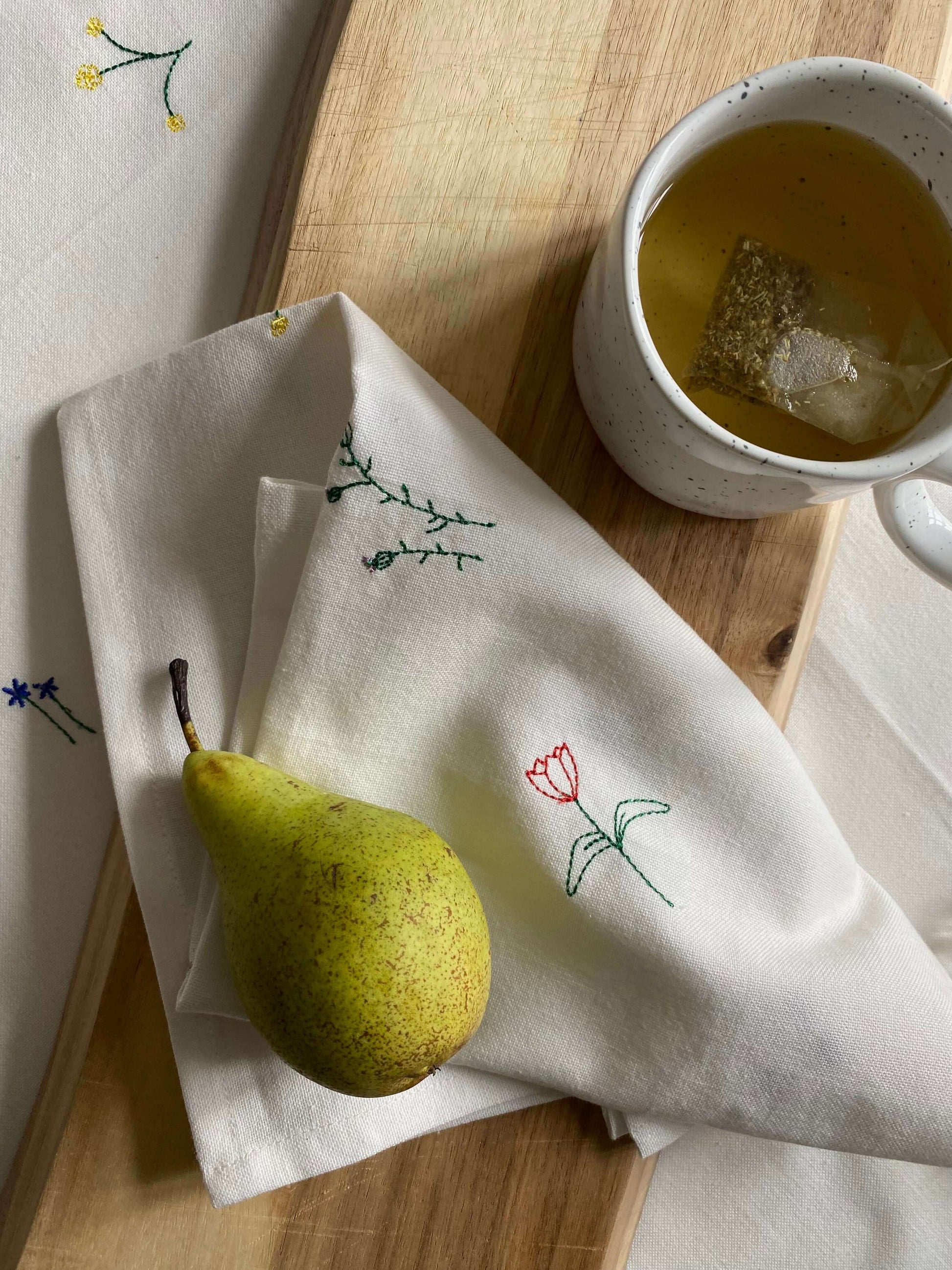 To hvide stofservietter med broderede blomster, placeret på et træbræt sammen med en pære.