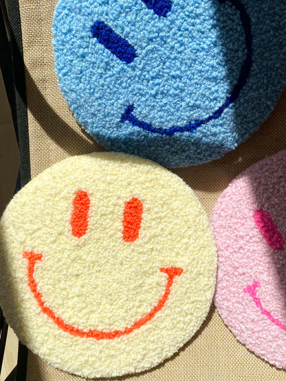 Tre Smiley vægtæpper i gul, blå og lyserød – lagt sammen på tekstiloverflade med sollys.