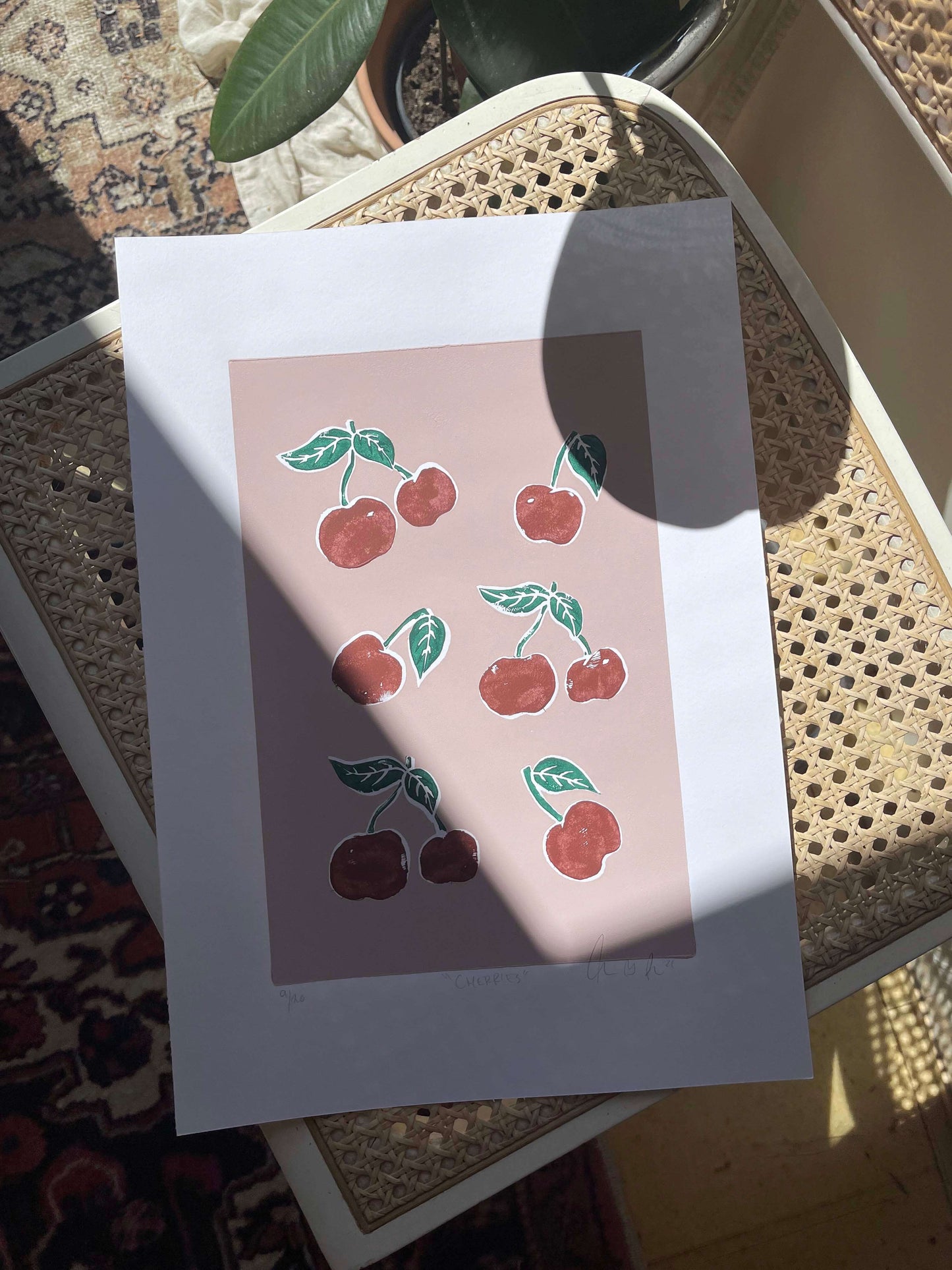 CHERRIES linoleumstryk med røde kirsebær i ramme, smukt opstillet på en stol.