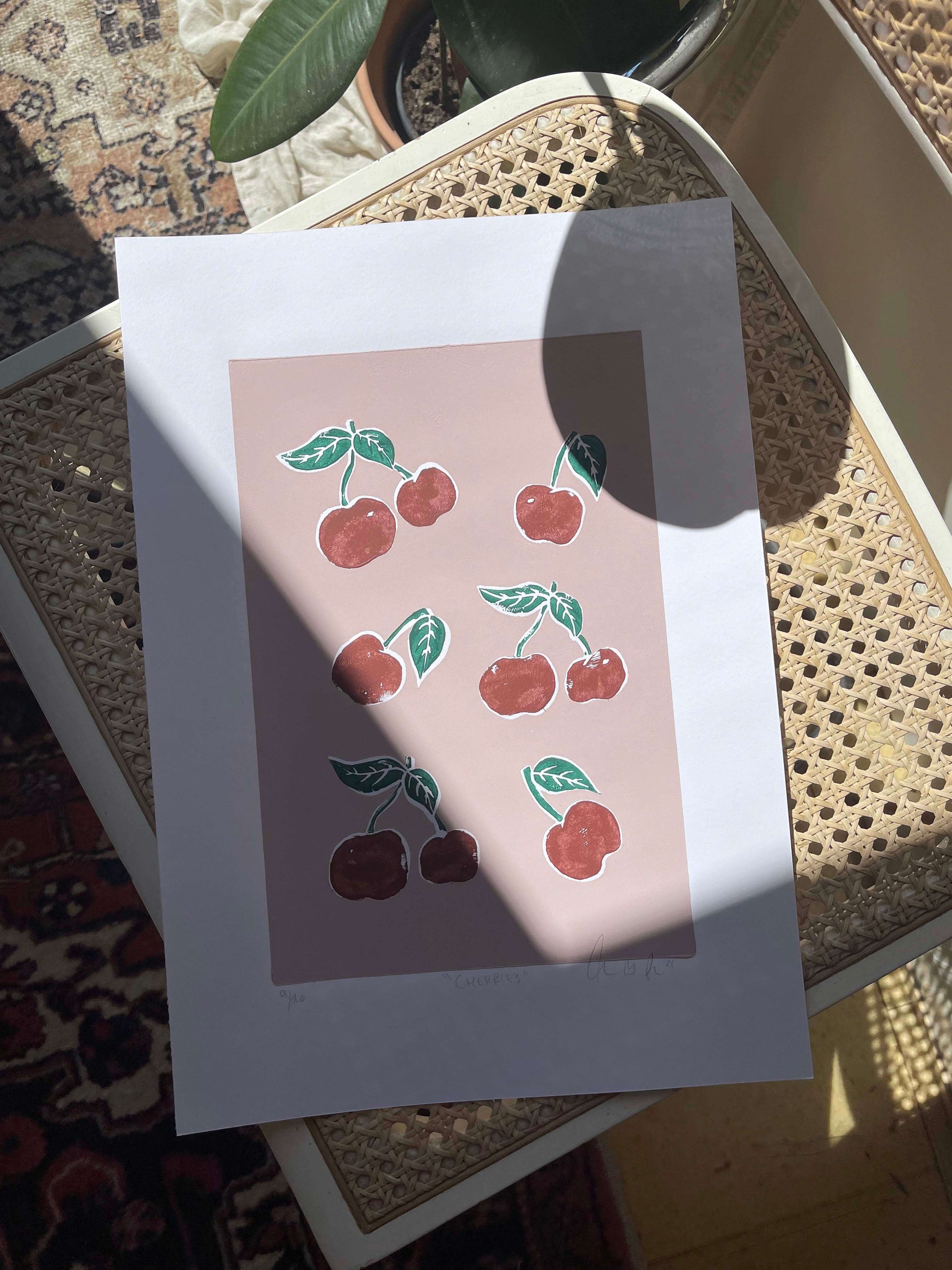 CHERRIES linoleumstryk med røde kirsebær i ramme, smukt opstillet på en stol.