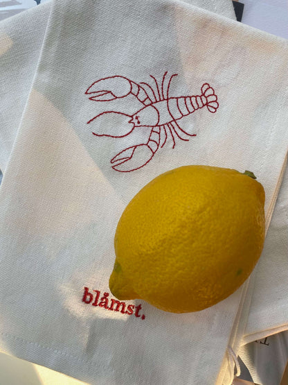 Stofserviet med rød hummerdetalje og Blåmst-logo i hjørnet